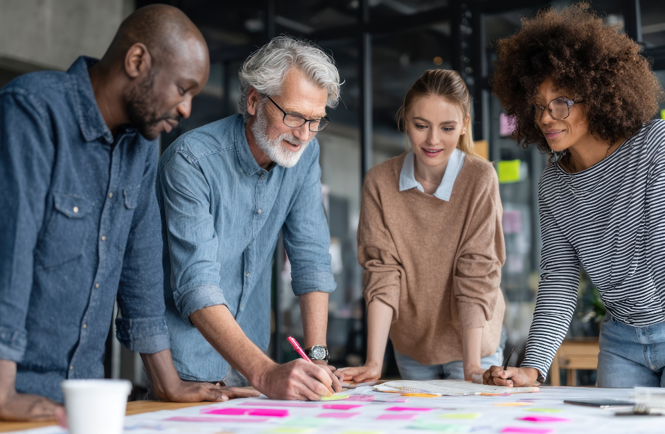 Four diverse professionals from different generations collaborate around a table covered in colorful sticky notes and documents, engaged in a creative planning session in a modern office space.