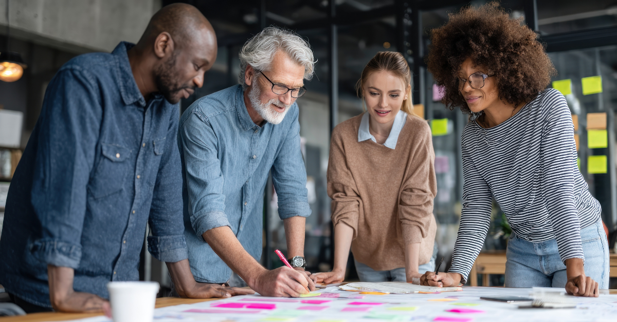 Four diverse professionals from different generations collaborate around a table covered in colorful sticky notes and documents, engaged in a creative planning session in a modern office space.