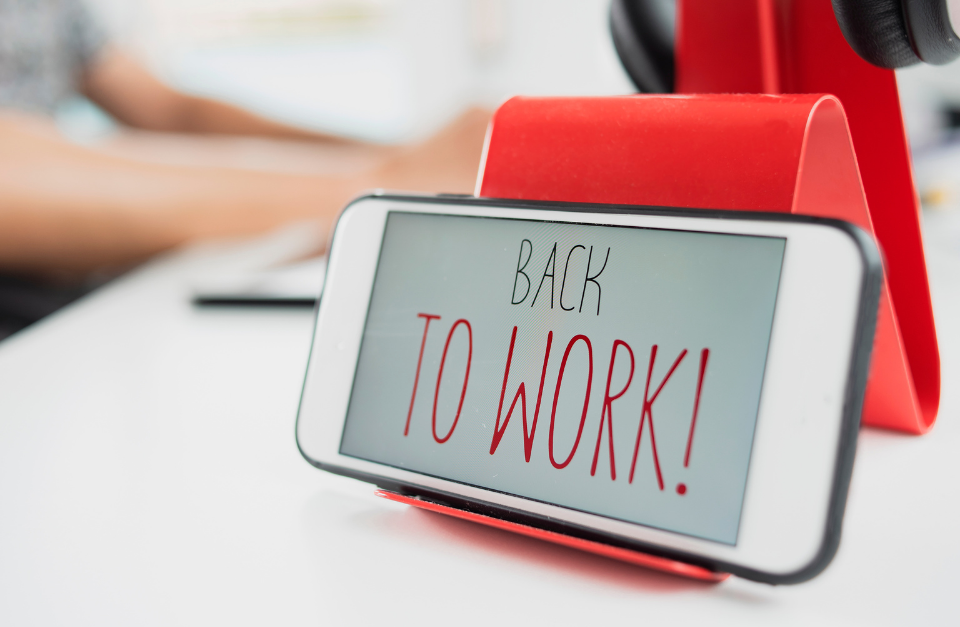 A smartphone on a red stand displaying the words "Back to Work!" on its screen, sitting on a white desk with blurred hands typing on a keyboard in the background.