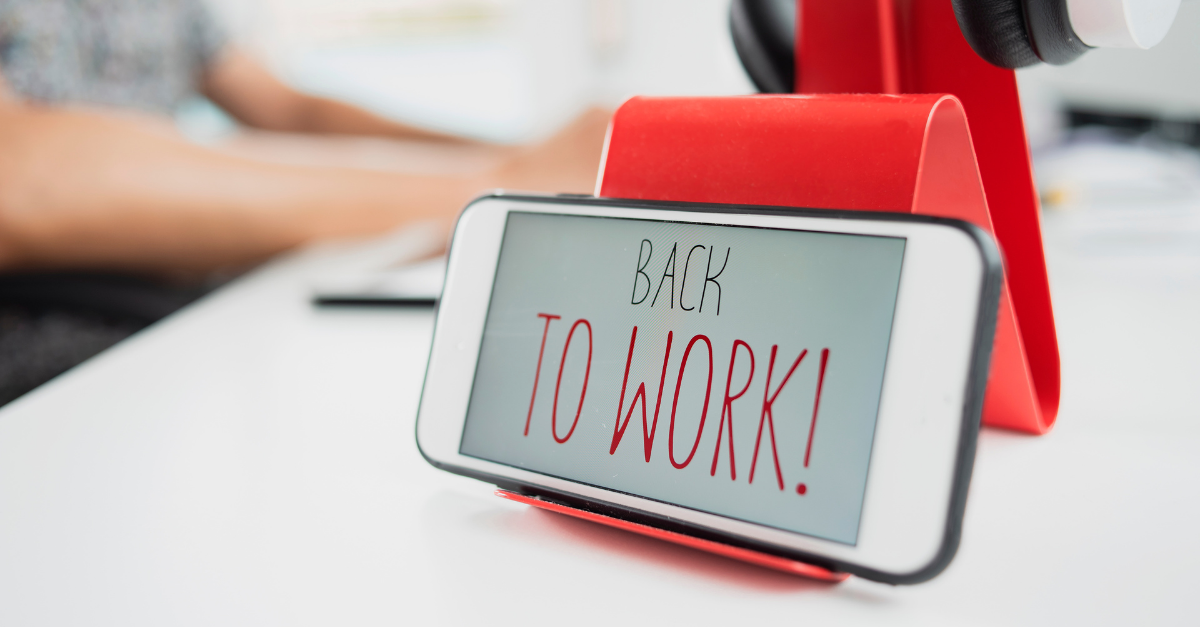A smartphone on a red stand displaying the words "Back to Work!" on its screen, sitting on a white desk with blurred hands typing on a keyboard in the background.
