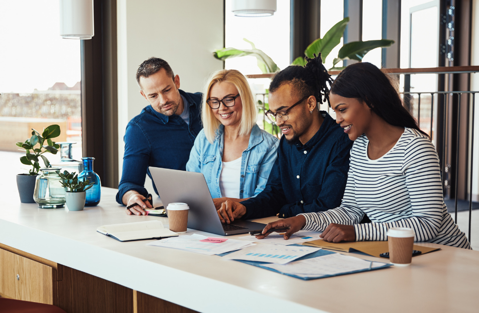 A diverse group of four professionals gathered around a laptop in a bright, modern office, collaborating on documents and charts.