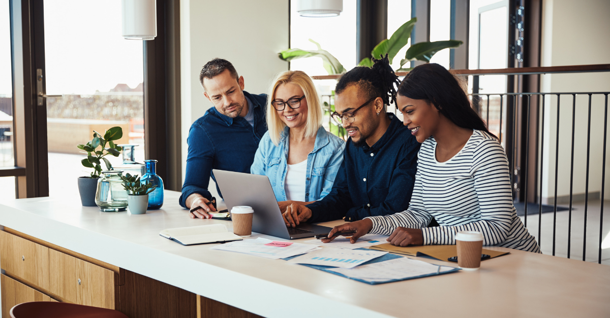 A diverse group of four professionals gathered around a laptop in a bright, modern office, collaborating on documents and charts.