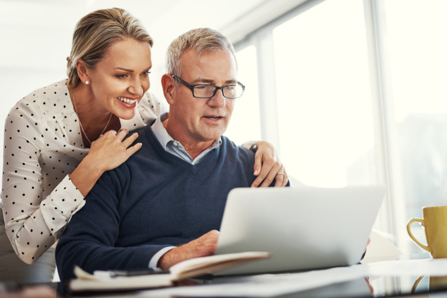 A middle-aged couple sits at a desk reviewing information on a laptop. The man, wearing glasses and a sweater, looks at the screen while the woman stands beside him with her hand on his shoulder, smiling. A notebook and a coffee mug sit on the desk in a bright, modern home setting.